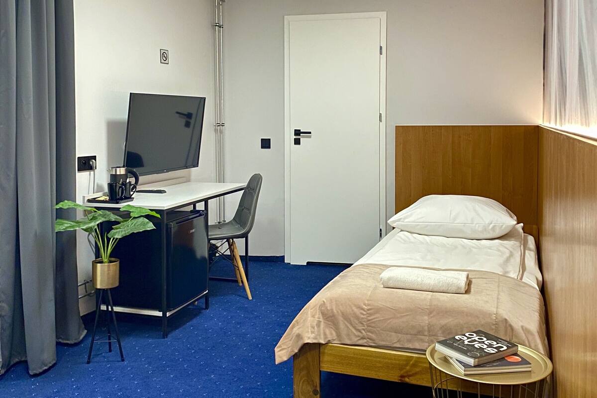 A hotel room with a single bed, a desk with a computer and a chair, a small table with a book, and a potted plant.