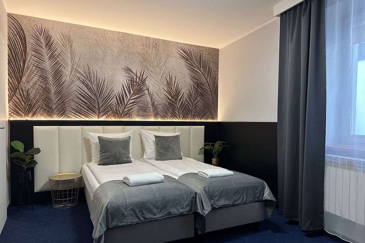 A modern hotel room with a large bed, two bedside tables, a wall-mounted light panel, and a view of the outdoors through a window with curtains.