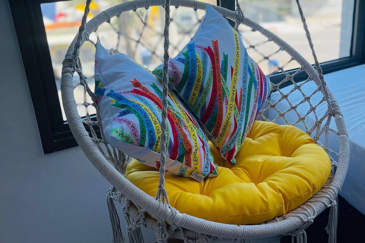 Una silla colgante de macramé con almohadones de colores y un cojín amarillo junto a una ventana con vista al mar.