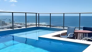 Una piscina en la azotea con vistas despejadas al océano, una silla de descanso blanca y una pequeña mesa.