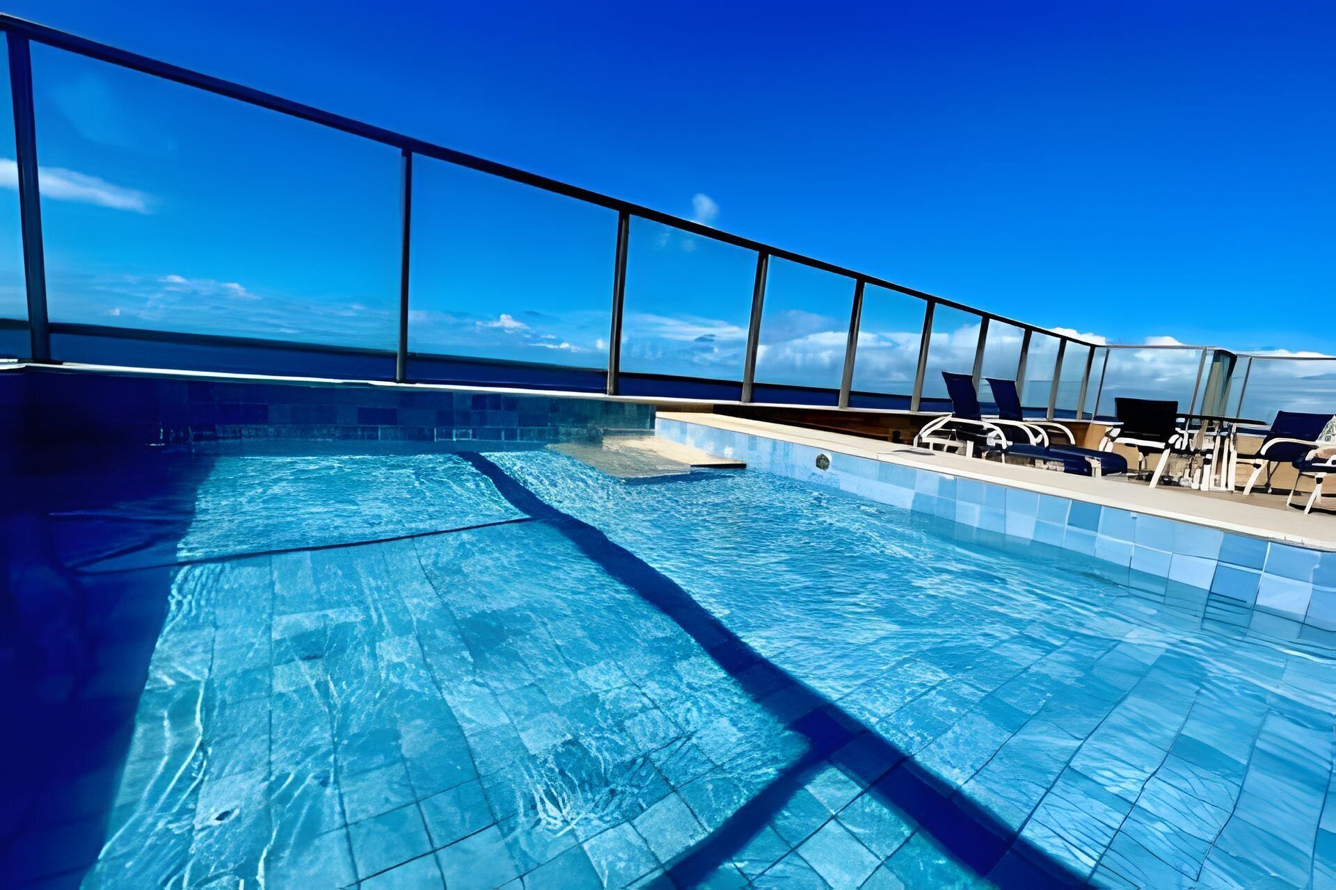 Une piscine à l’eau d’un bleu cristallin, entourée de chaises longues et bordée d’une rambarde en verre.