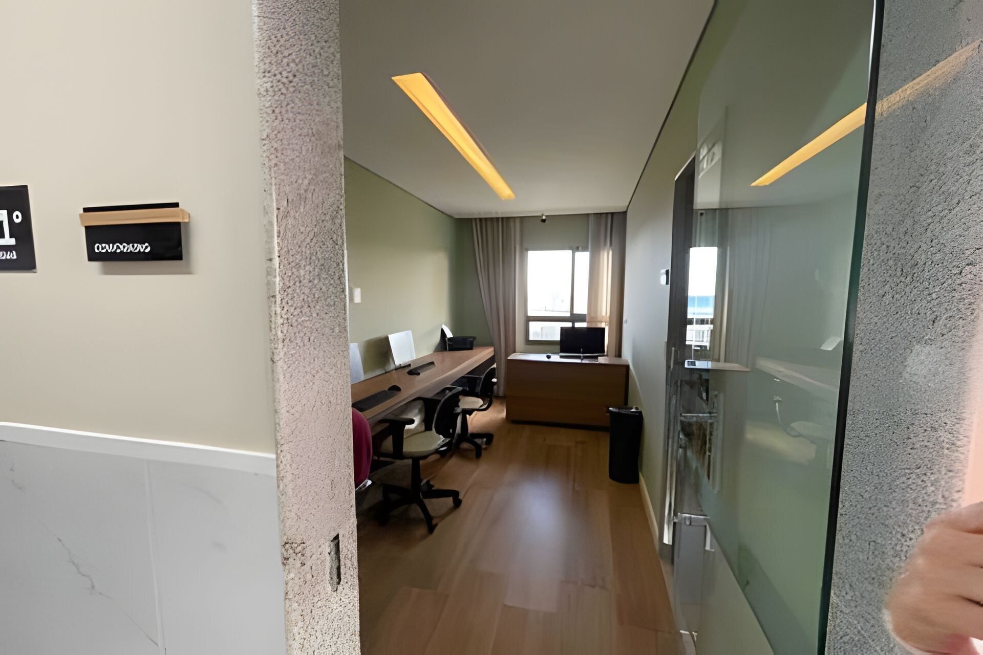 Quarto de hotel moderno com divisória de vidro, escrivaninha com computador, cadeira e uma janela grande.