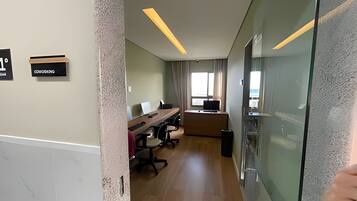 Quarto de hotel moderno com divisória de vidro, escrivaninha com computador, cadeira e uma janela grande.