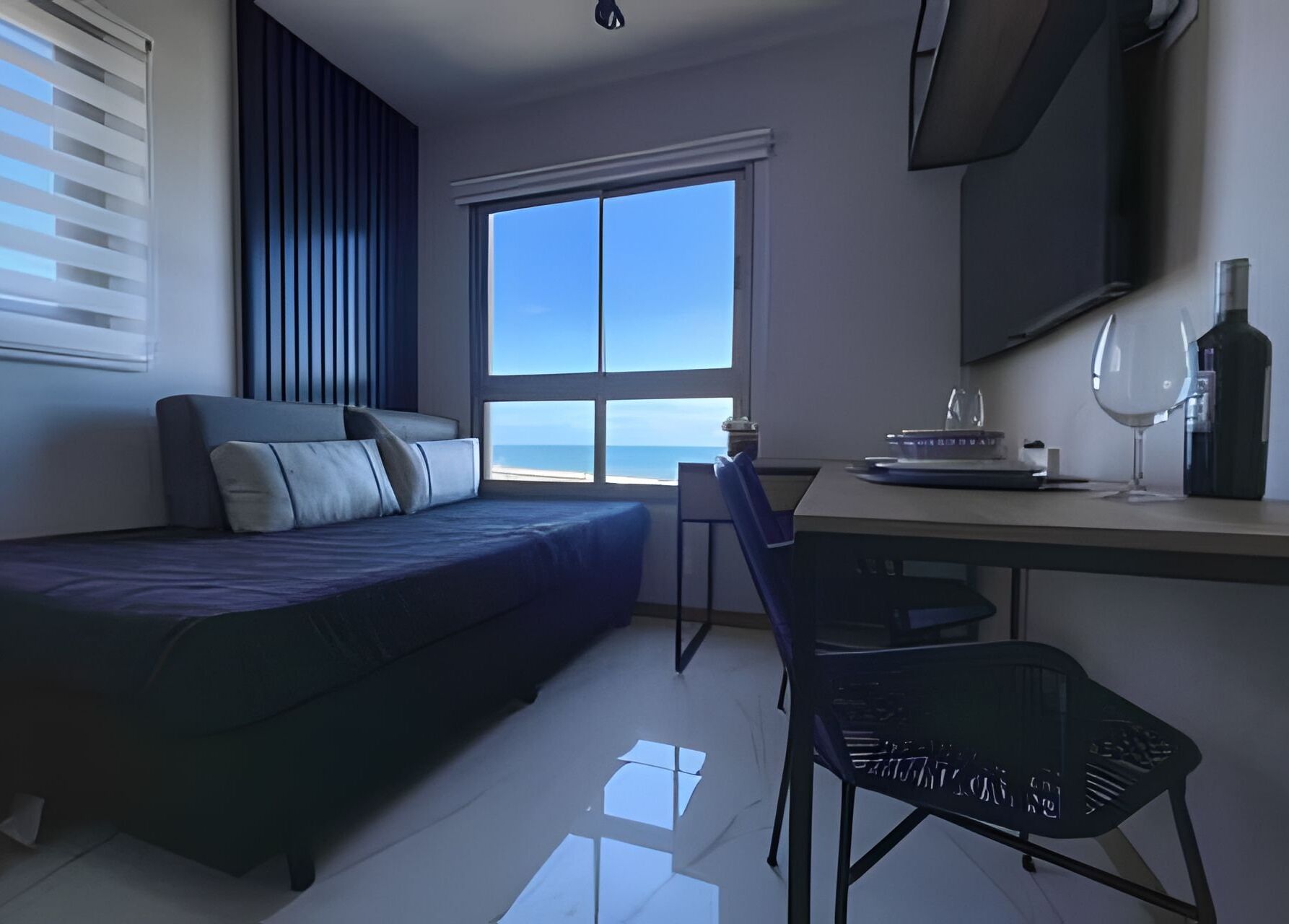 A modern bedroom with a bed, desk, chair, and a large window offering a view of the ocean.