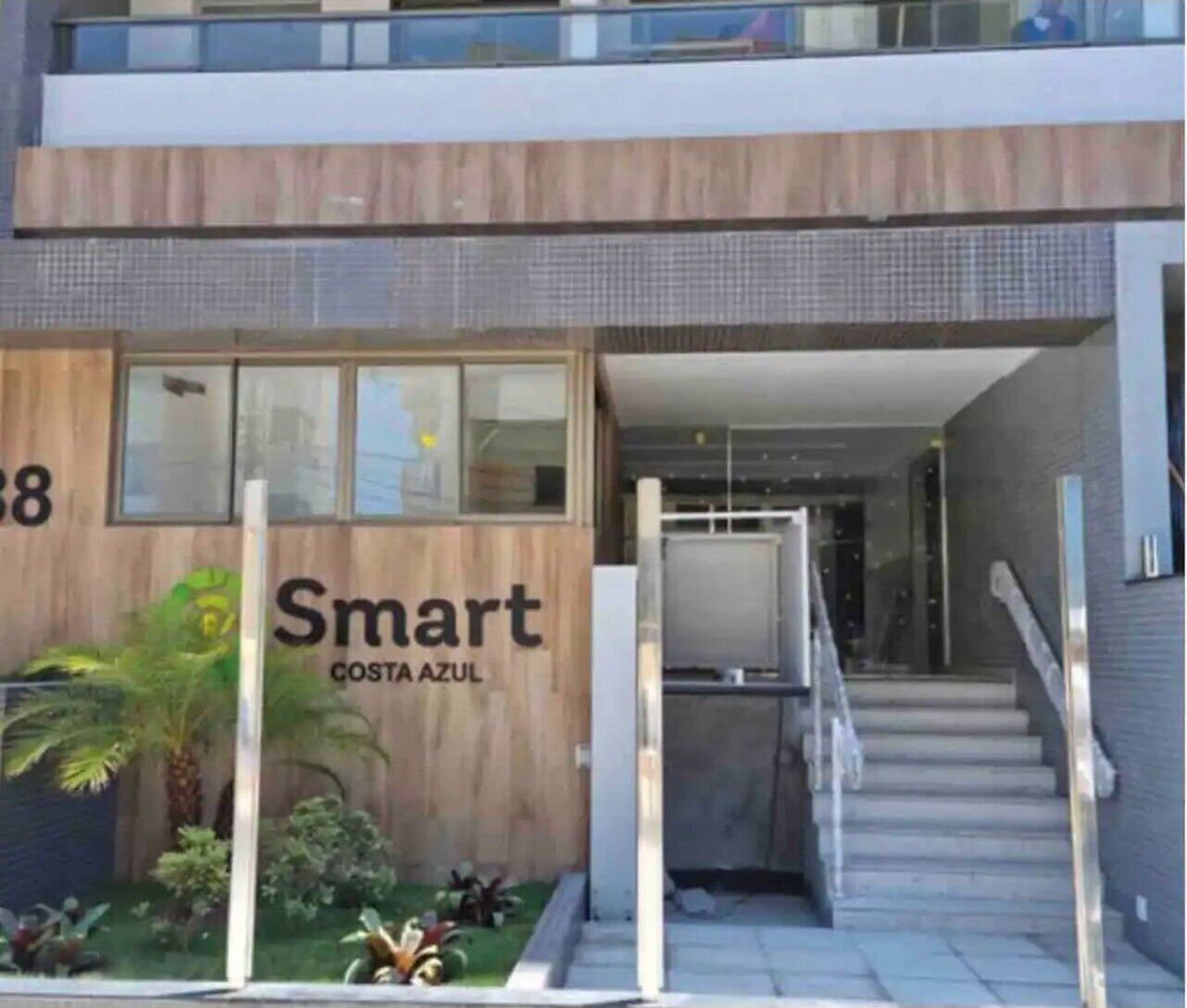 Um edifício moderno com fachada de madeira e a inscrição "Smart Costa Azul" na frente. Há degraus que levam à entrada, e uma pequena área de jardim com plantas e uma palmeira.