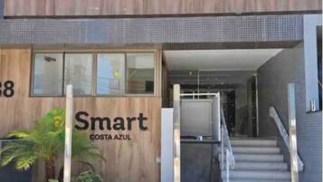 Um edifício moderno com fachada de madeira e a inscrição "Smart Costa Azul" na frente. Há degraus que levam à entrada, e uma pequena área de jardim com plantas e uma palmeira.