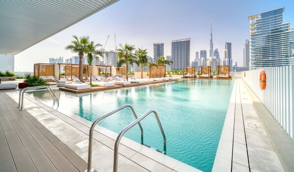 A rooftop infinity pool with lounge chairs and palm trees, overlooking a city skyline with modern buildings. - Prime Location Pristine Studio in Business Bay (Dubai)