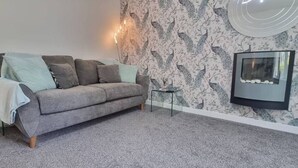 A living room with a grey sofa, a glass coffee table, a lamp, and a fireplace with a framed picture. - The Pennington Apartment Leigh (Leigh)