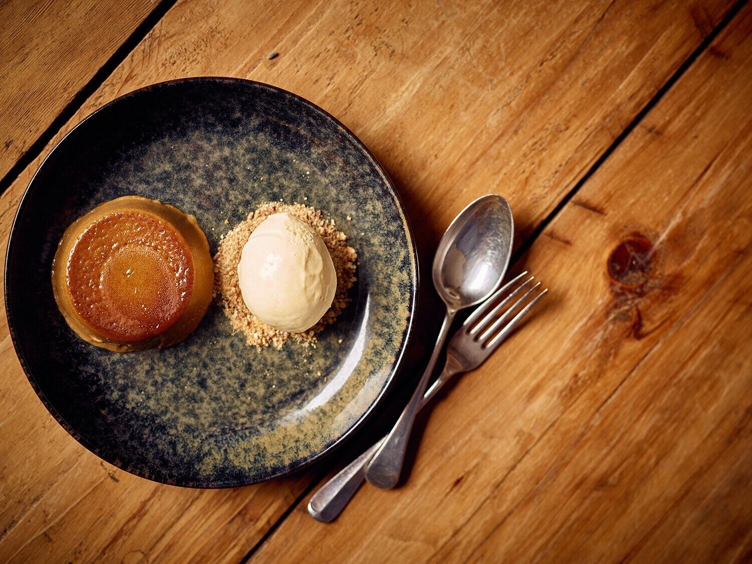 A dessert plate with a caramel and ice cream on a dark ceramic plate, accompanied by a fork and spoon on a wooden table.