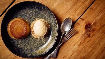 A dessert plate with a caramel and ice cream on a dark ceramic plate, accompanied by a fork and spoon on a wooden table.