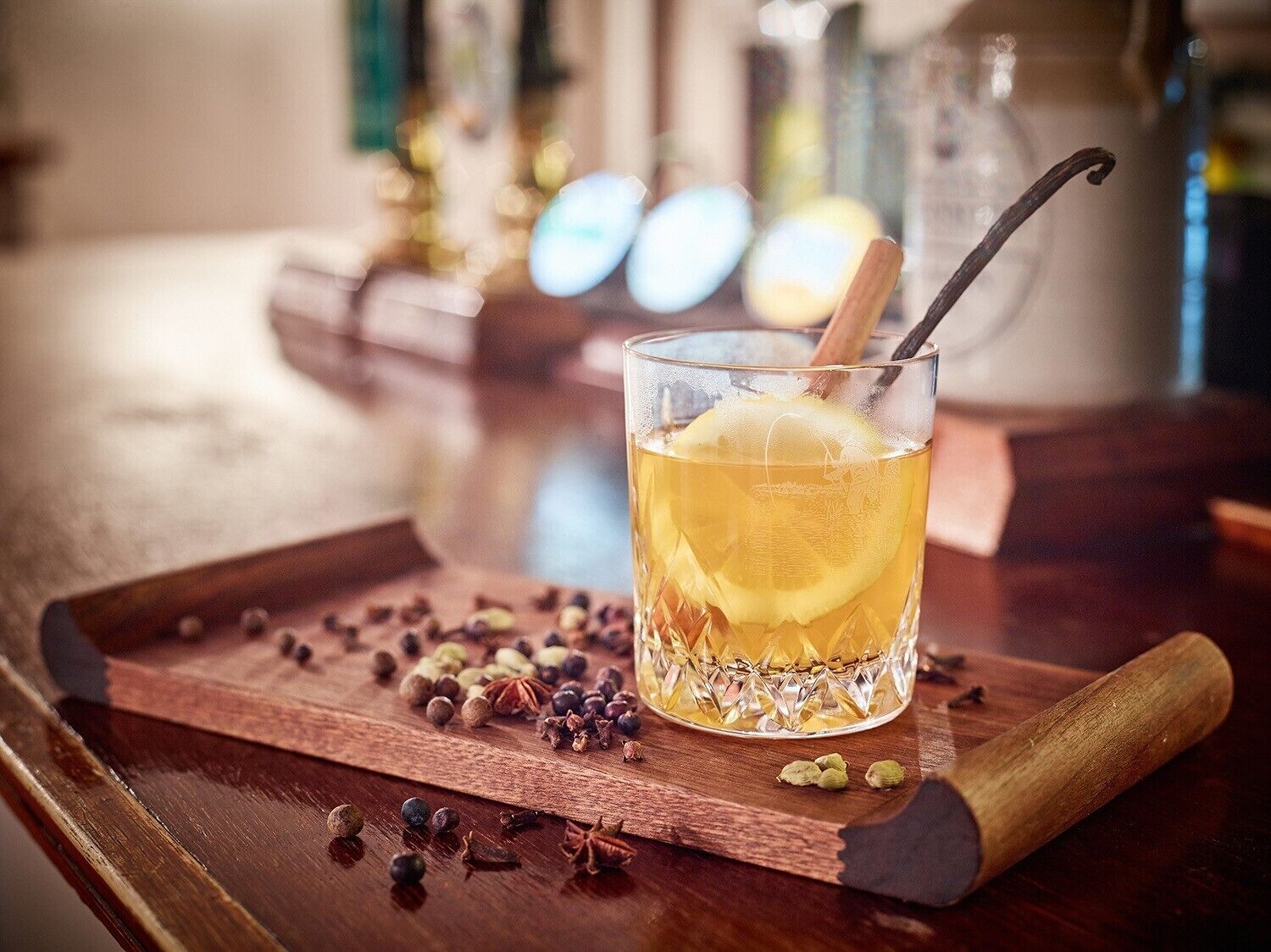 A glass of amber liquid with a lemon slice and a cinnamon stick, surrounded by scattered spices on a wooden tray.