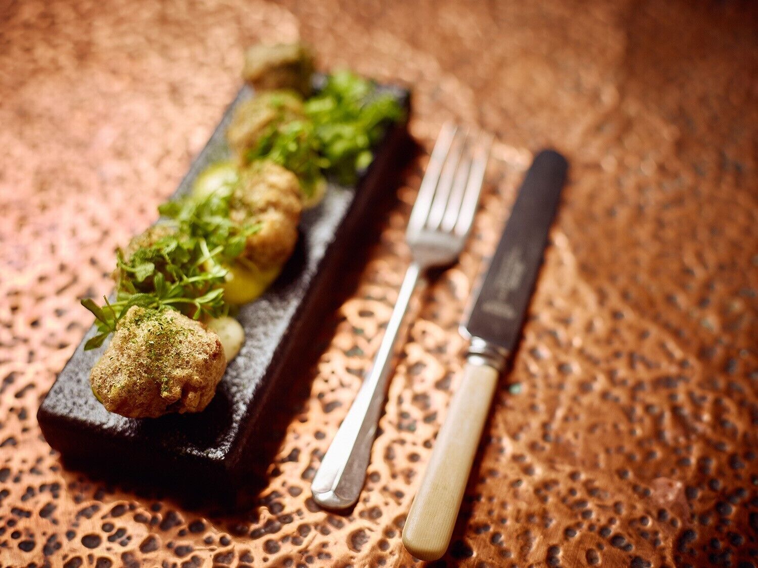 A rectangular black slate serving dish with a series of golden-brown fried items and green garnish, accompanied by a fork and knife on a textured surface.