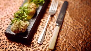 A rectangular black slate serving dish with a series of golden-brown fried items and green garnish, accompanied by a fork and knife on a textured surface.