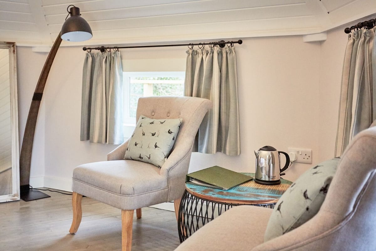 A cozy room with a light grey armchair, a round wooden side table with a kettle and a notebook, and a floor lamp.