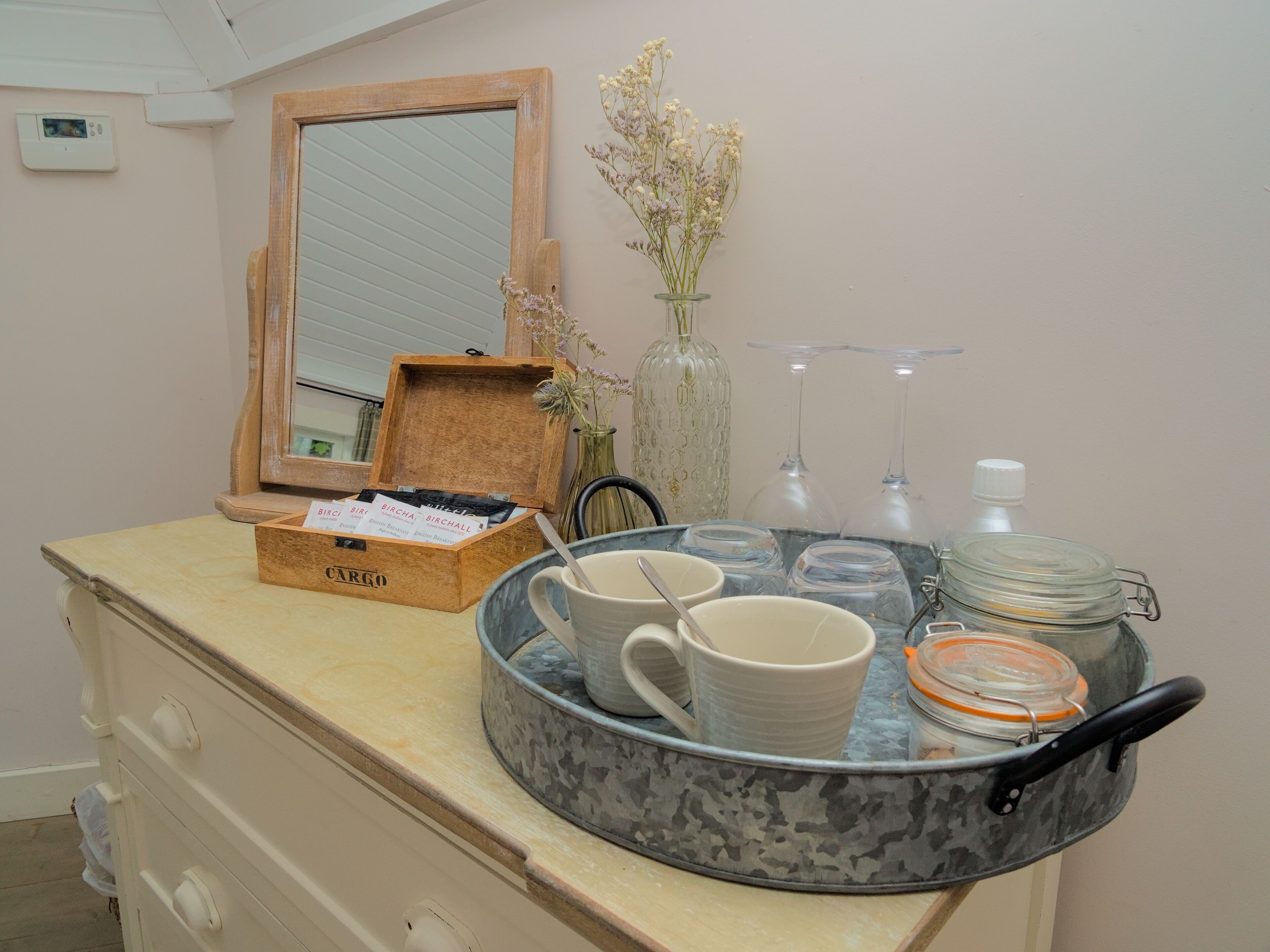 Una encimera de cocina con una caja de madera que dice "CARGO", una bandeja con tazas y un tarro de cristal, y un jarrón con flores.