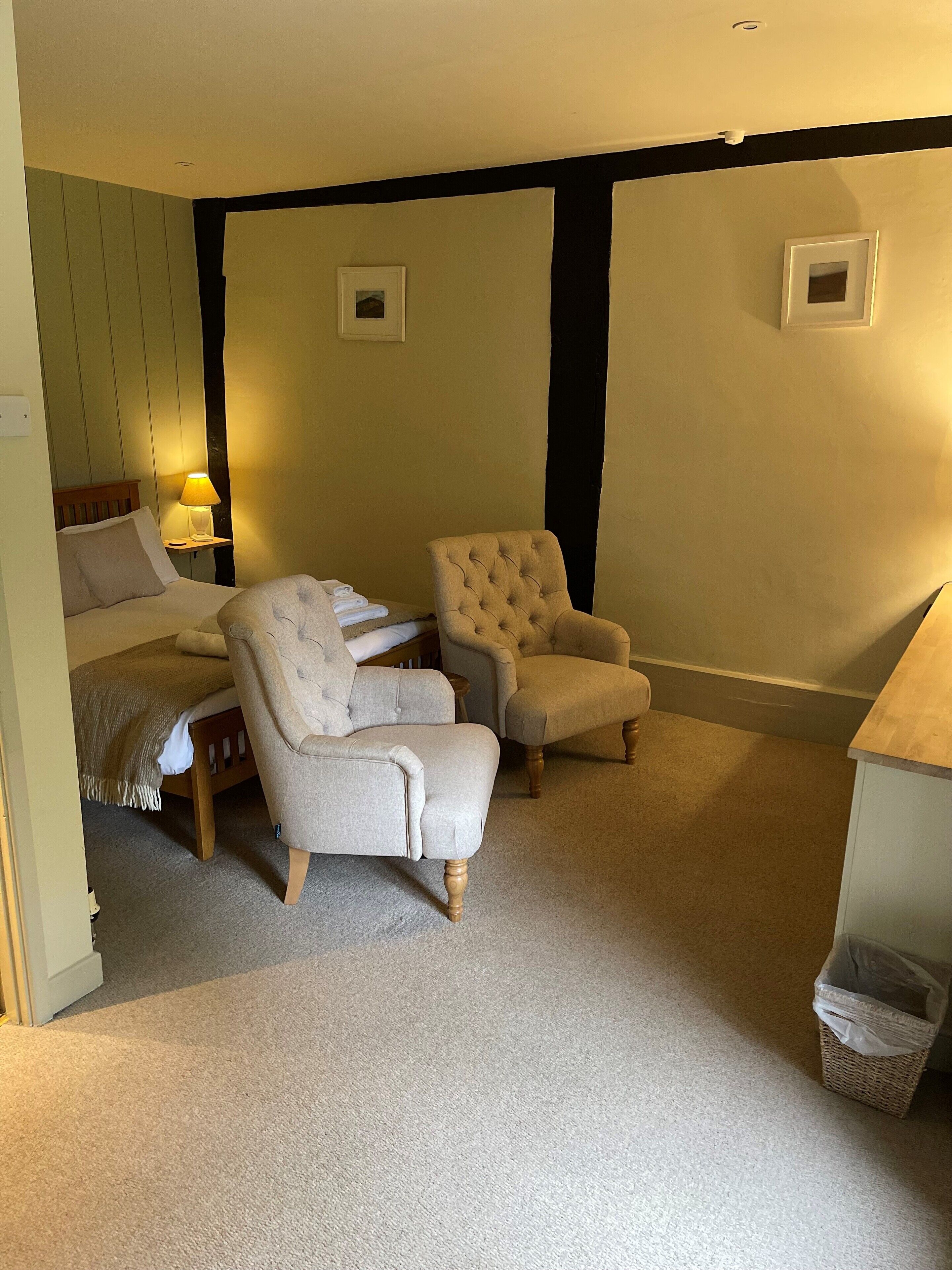 A bedroom with a bed, two armchairs, a bedside table with a lamp, and a small trash bin.
