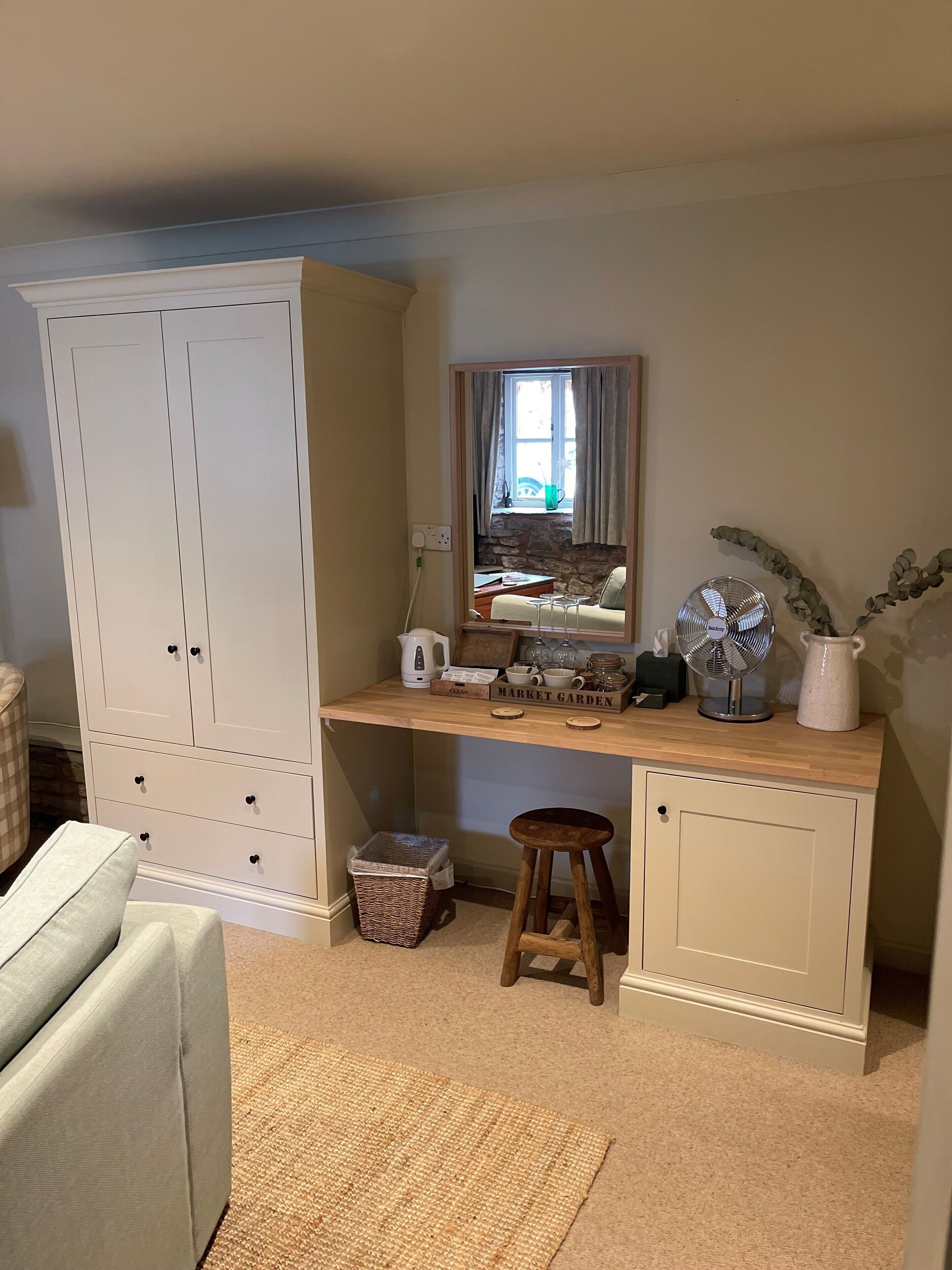 Een kamer met een grote witte garderobe, een houten bureau met een spiegel, een klein krukje en een rieten mand.