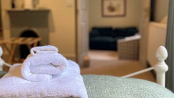 A neatly made bed with folded towels, a green textured bedspread, and a view of a living area with a sofa and a fireplace in the background.