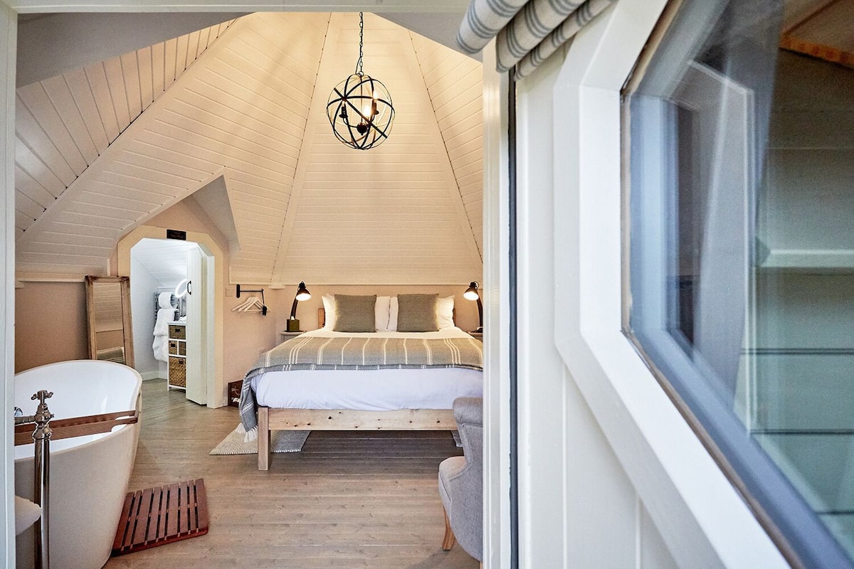A bedroom with a sloped ceiling, a bed with a striped bedspread, bedside lamps, and a hanging light fixture.