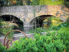 Un puente de piedra sobre un río tranquilo, rodeado de abundante vegetación.