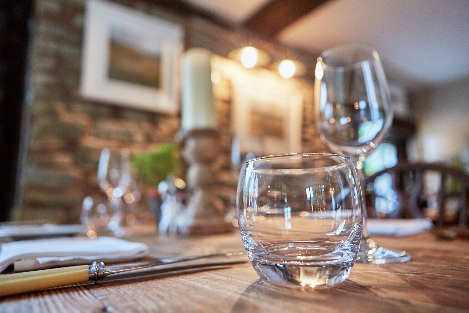A dining table with a clear glass, a pen, and a candle in a cozy, warmly lit room with a brick wall and framed picture.