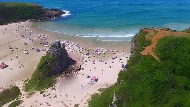 Vista aerea di una spiaggia con numerosi ombrelloni e persone, circondata da vegetazione e scogliere.