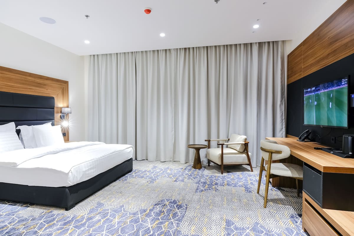A hotel room with a bed, a desk with a TV, a chair, and a large window with curtains.