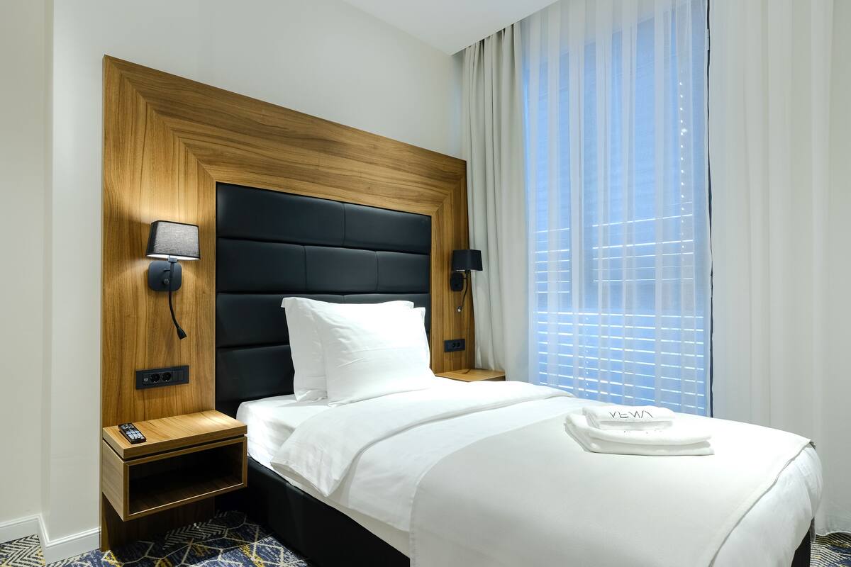 A hotel room with a wooden headboard, a white bedspread, two bedside tables with lamps, and a window with blinds.