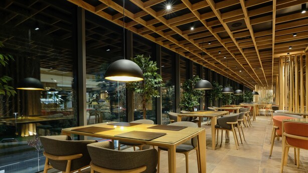 A modern restaurant interior with wooden tables and chairs, pendant lighting, and large windows offering views of the outdoors.