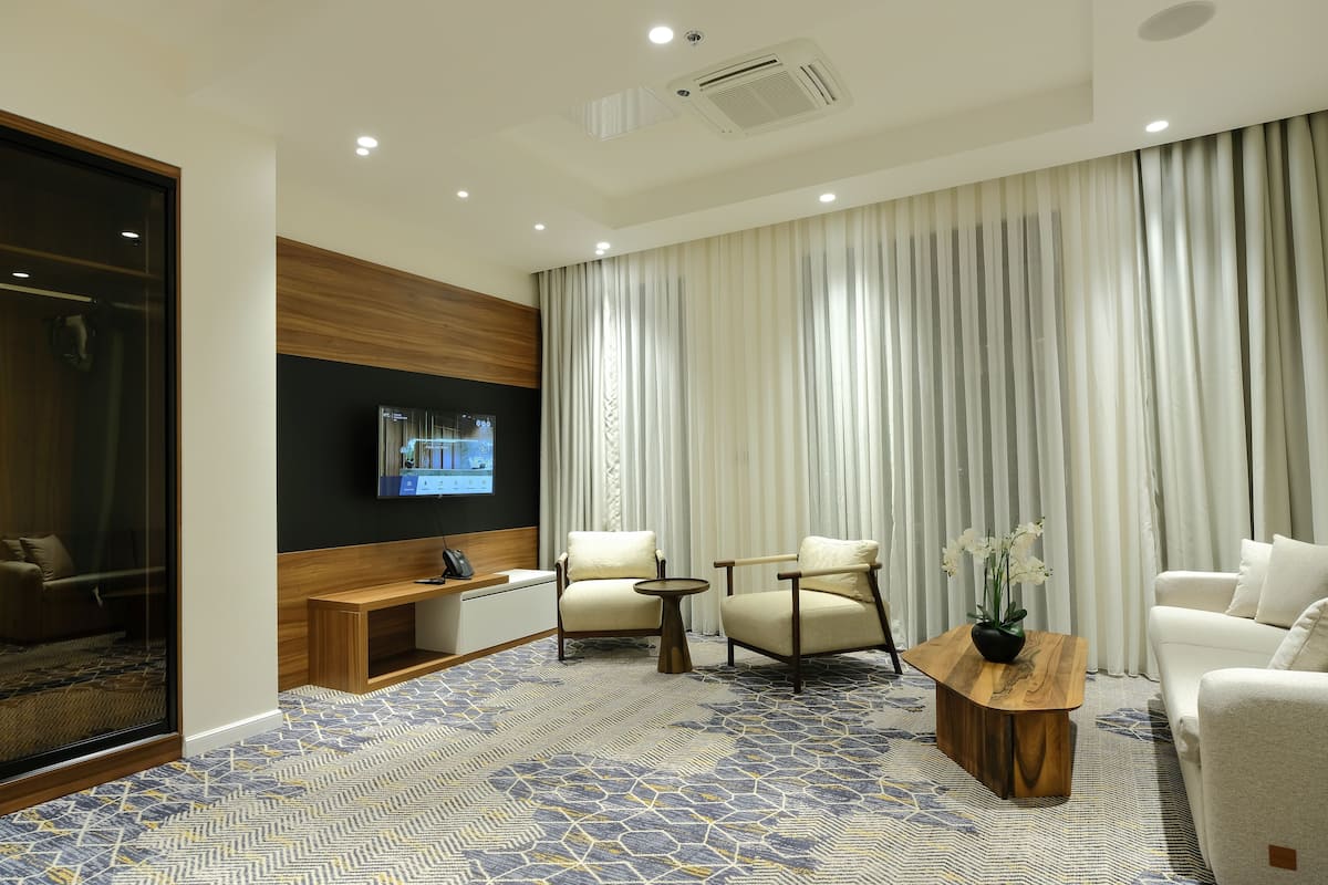 A modern hotel room with a large window, a flat-screen TV mounted on the wall, a wooden bench, two armchairs, a round coffee table, and a potted plant.