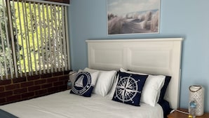 A bedroom with a large bed, a nightstand, a window with blinds, and a painting on the wall.
