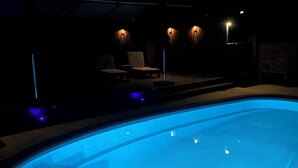 A swimming pool with a blue light, lounge chairs, and wall-mounted lights.