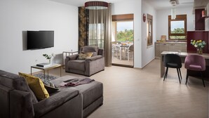 A modern living room with a sofa, armchair, and a flat-screen TV. There is a dining area with a table and chairs, and a kitchen area with a sink and cabinets. - Malibu Grand Royale - Luxury Group Villa with Pool (Vir)