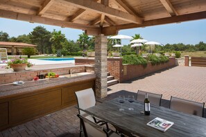 An outdoor patio with a wooden table, chairs, and a pool in the background. - Malibu Grand Royale - Luxury Group Villa with Pool (Vir)