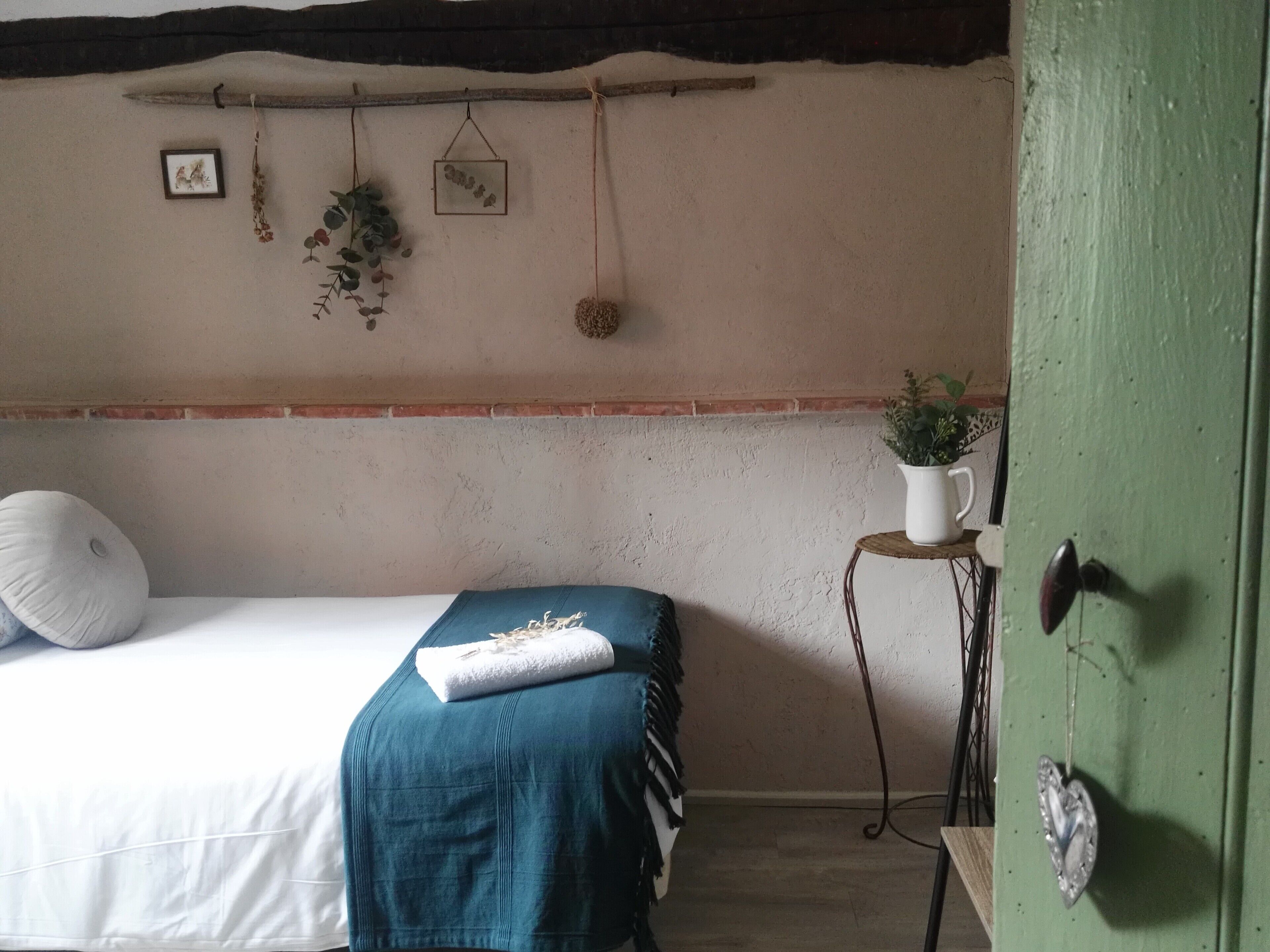 A bedroom with a bed, a pillow, a blue blanket, a small plant on a side table, and a wall-mounted shelf with decorative items.