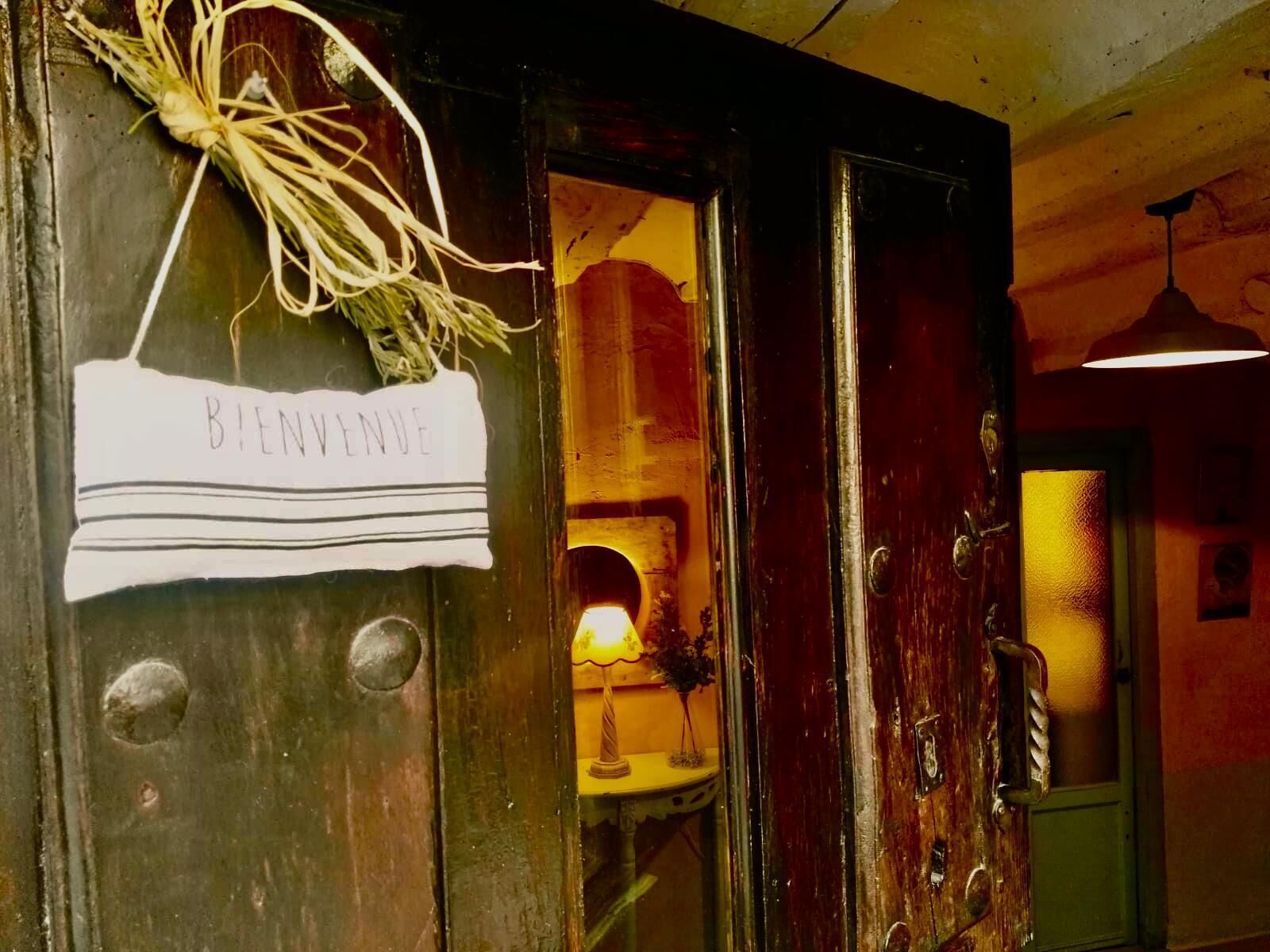 Uma porta de madeira com uma placa que diz 'BIENVENUE', uma planta pendurada e uma pequena mesa com abajur e flores no interior.
