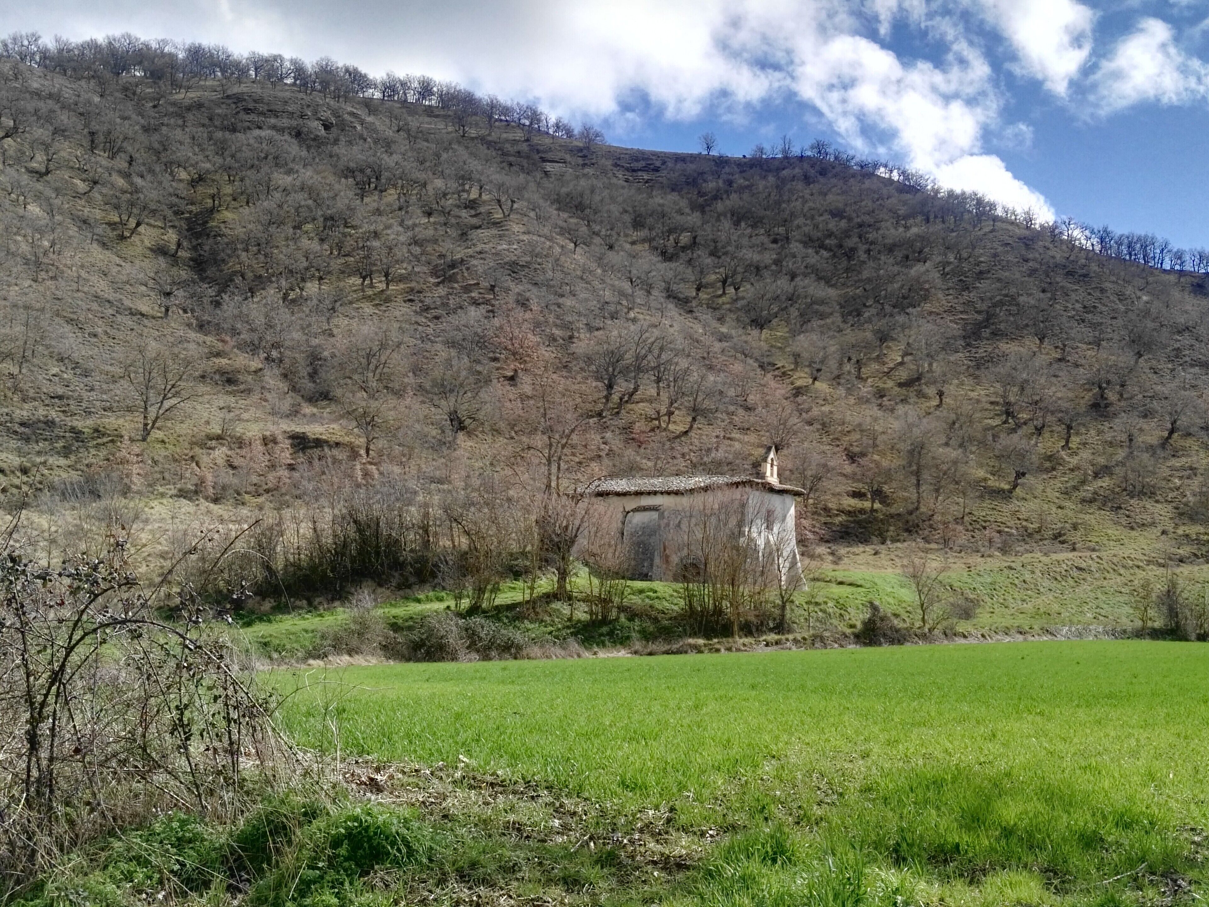 Paisaje rural con una pequeña construcción en una ladera, rodeada de campos y algunos árboles.