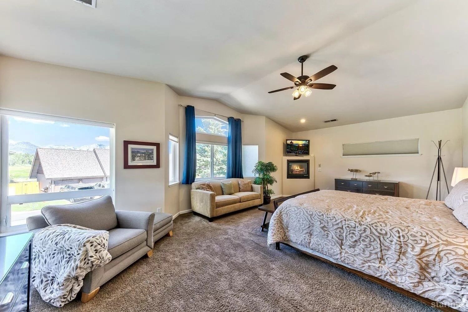 A bedroom with a large bed, a sofa, a chair, a ceiling fan, a fireplace, and a view of the outside.