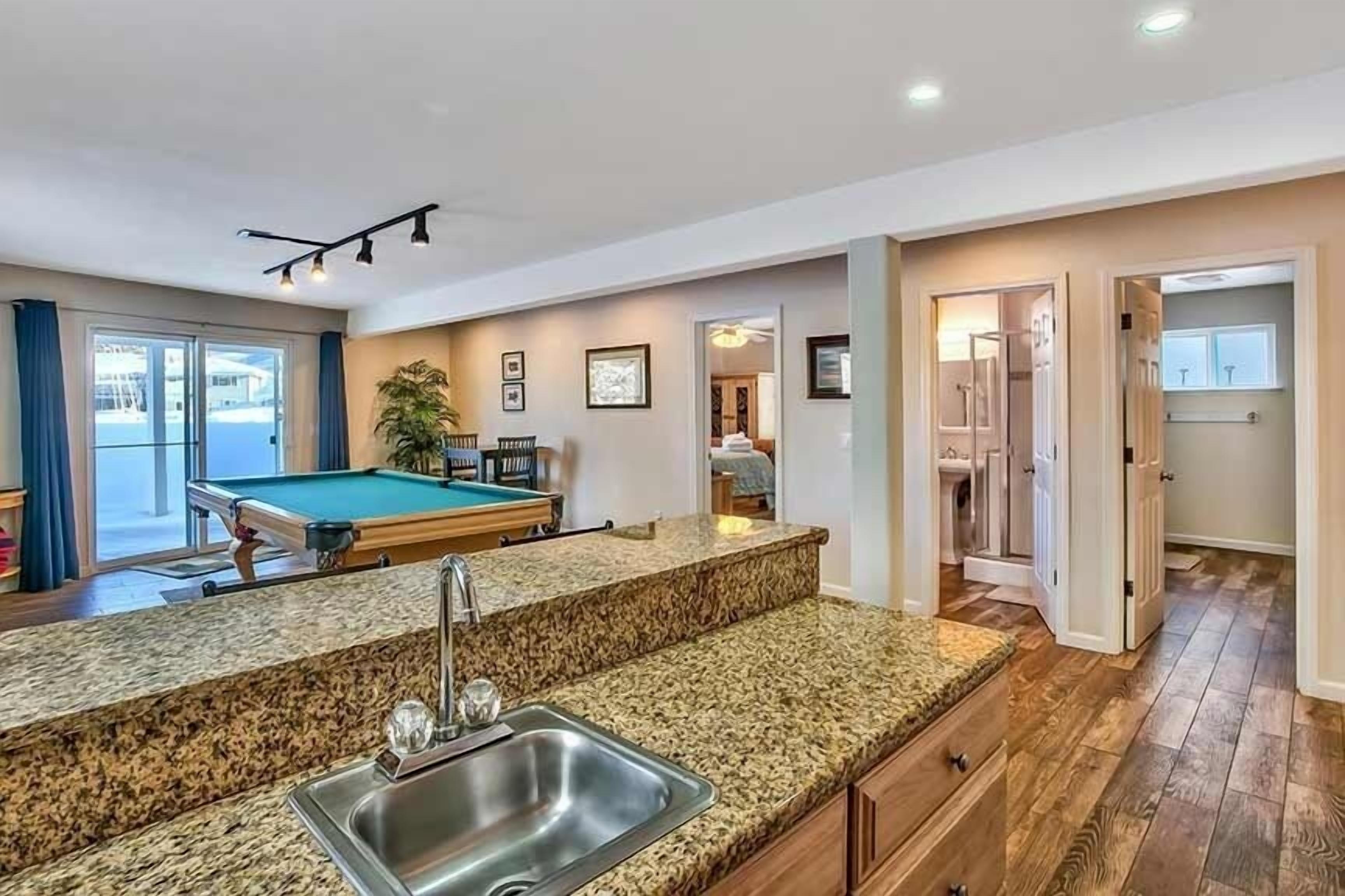 A modern kitchen with a granite countertop, stainless steel sink, and a pool table in the background.
