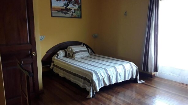 A bedroom with a wooden floor, a bed with striped bedding, a painting on the wall, and a window with curtains. - Hotel le Colbert (Antsiranana)
