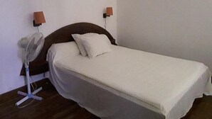 A single bed with a white mattress and pillows, a bedside table, a wall-mounted fan, and two wall-mounted lamps. - Hotel le Colbert (Antsiranana)