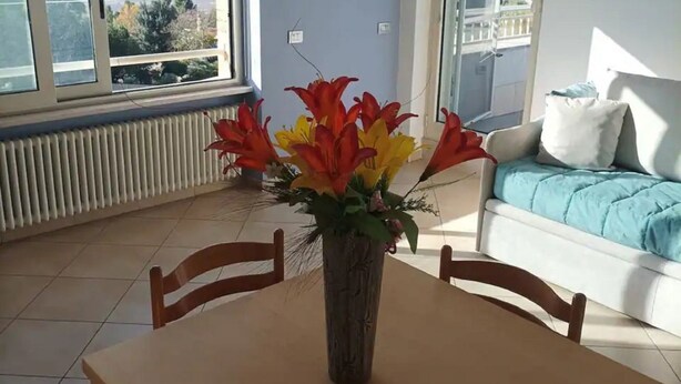 Un comedor con mesa y sillas, un florero con flores, un sofá con una manta azul y vista al mar a través de amplias ventanas.