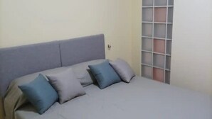 Una cama con cabecera gris y dos almohadas, una ventana con diseño de cuadrícula y una pared lisa.