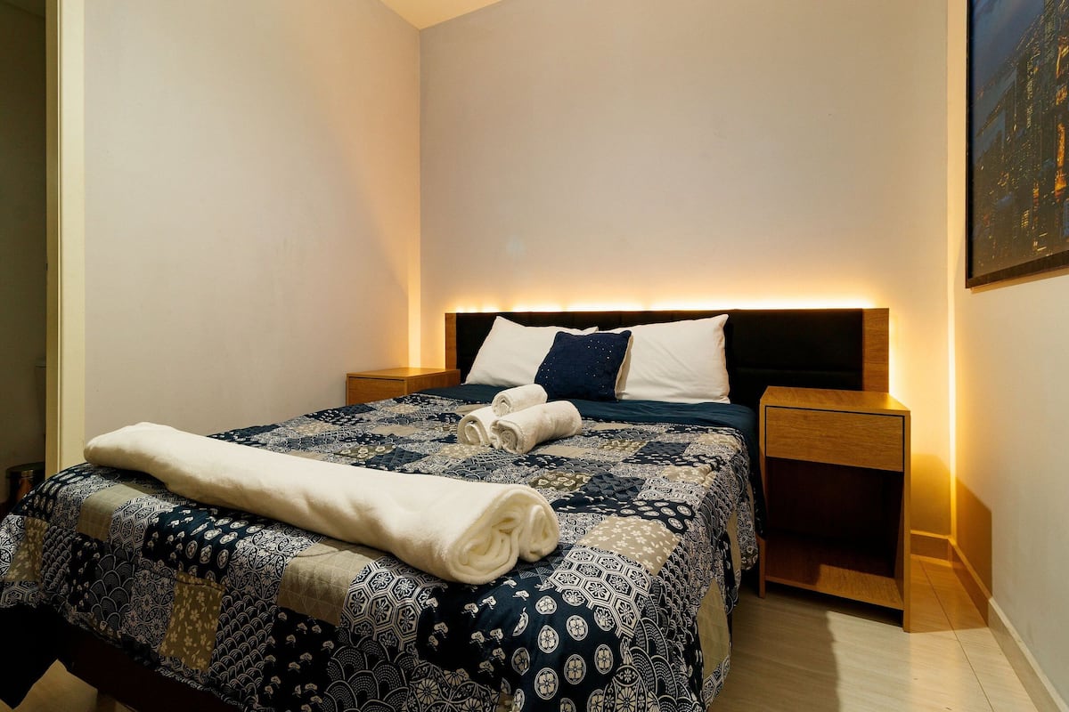 A neatly made bed with a patterned bedspread and rolled towels, a wooden nightstand, and a wall-mounted lamp.