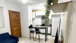 Un ambiente compatto con angolo cottura, zona pranzo con sgabelli, divano blu e ventilatore a soffitto.