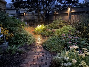 A garden pathway with a brick surface, surrounded by various plants and flowers.