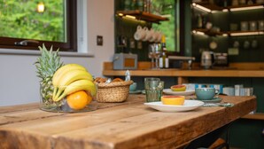 Una mesa de madera con frutas, un vaso y una taza, con una cocina y una ventana al fondo.