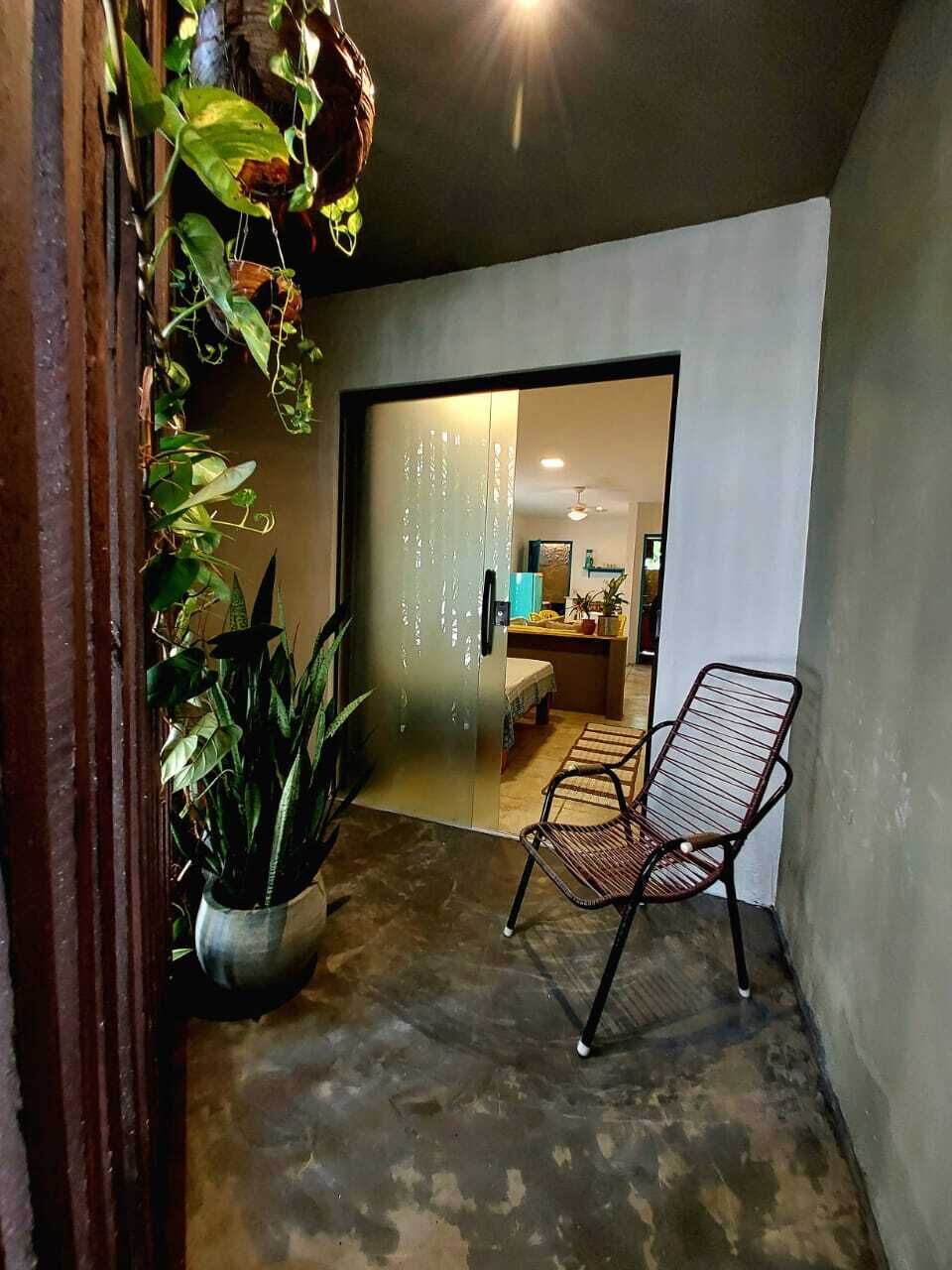 A hallway with a chair, a potted plant, and a doorway leading to a room with a bed and a table.