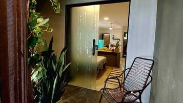 A hallway with a chair, a potted plant, and a doorway leading to a room with a bed and a table.