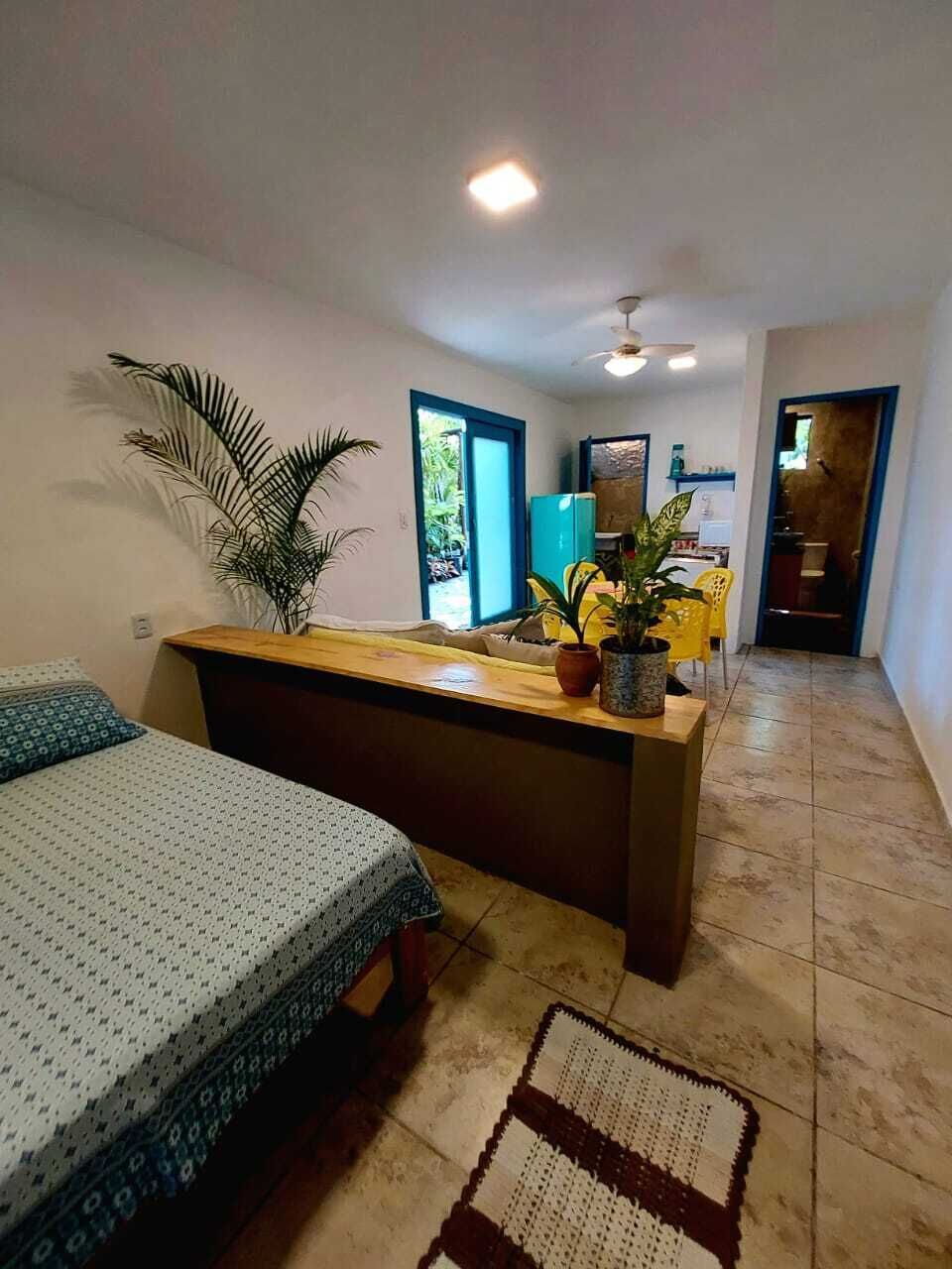 A hotel room with a bed, a desk, a potted plant, and a view of the kitchen area.